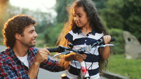 Closeup.-Portrait-of-a-little-girl-and-her-dad-next-to-the-bike.-Dad-hugs-her.-Looking-at-each-other,-then---into-the-camera.-Smiling.-Blurred-background